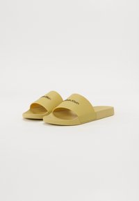 Pantofi de plajă galbeni din cauciuc. Au o curea lată cu logo-ul "Calvin Klein". Textură netedă, talpă plată, design cu vârf deschis.