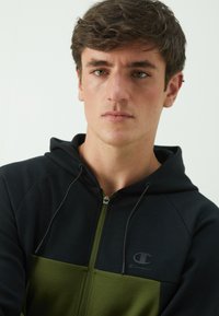 Kapuzensweatjacke mit Reißverschluss, die oben schwarz und unten olivgrün ist. Weicher Stoff mit kontrastierenden Farbblöcken und Logodetail auf der Brust.