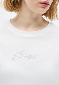 Κοντινή λήψη ενός ατόμου που φοράει λευκό πουκάμισο με μικρές στρας που σχηματίζουν την καλλιγραφική λέξη «Softe» στο στήθος.