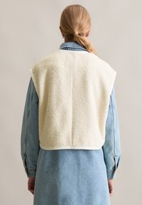 Kremfarget, teksturert fleecevest med avrundet kant, iført over en denimskjorte. Har ermeløs design og kontrastfôr.