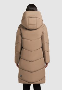 Kvinde iført en lang, beige polstret vinterjakke med høj krave og hætte, stående med ryggen til en ensfarvet baggrund.