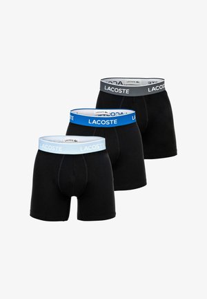 Three black boxer briefs with Lacoste branded elastic waistbands in light blue, blue, and dark gray, arranged in a staggered row.