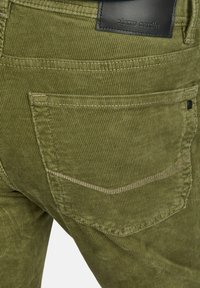 Grüne Cordhosen mit gerippter Textur, ausgestattet mit einer Gesäßtasche und einem schwarzen Lederetikett. Gestickte Akzente in einer helleren Farbe.