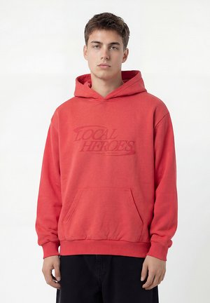 Młody mężczyzna w czerwonej bluzie z kapturem i przednią kieszenią, z napisem "Local Heroes", stojący na jednolitym białym tle.