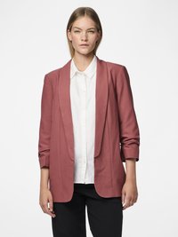 Jeune femme portant un blazer rose poussiéreux par-dessus une chemise blanche boutonnée et un pantalon noir, debout devant un fond blanc uni.