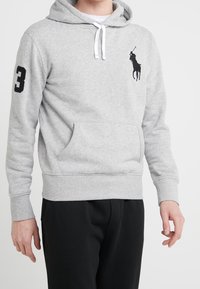 Sudadera gris de mezcla de algodón, con un bolsillo tipo canguro, puños elásticos y un logo de caballo negro en el pecho. El modelo lleva pantalones negros.
