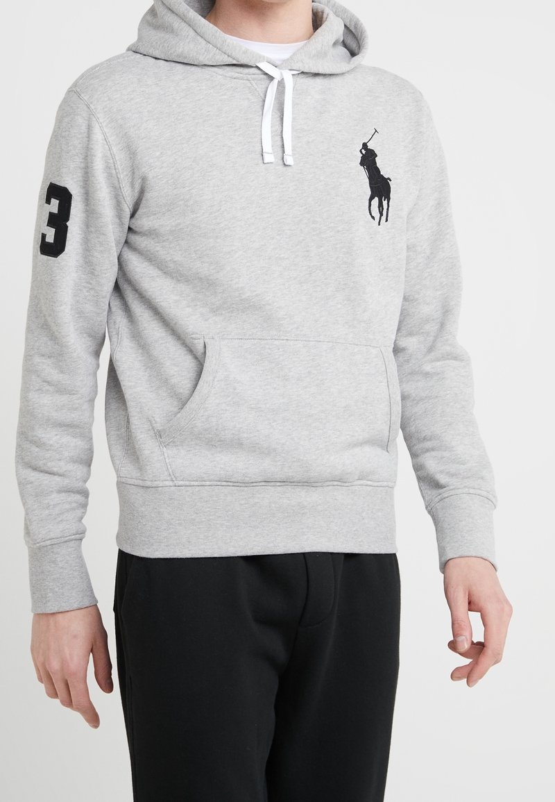 Sudadera gris de mezcla de algodón, con un bolsillo tipo canguro, puños elásticos y un logo de caballo negro en el pecho. El modelo lleva pantalones negros.