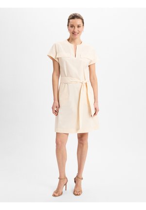 Licht perzikkleurige jurk met een V-hals, korte mouwen en twee voorzakken. De jurk heeft een taille met een riem en een rechte snit.