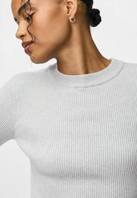 Gaiši pelēks ribots adījums ar apaļu kakla izgriezumu un garām piedurknēm. Tuvplāns uzsver faktūru un auduma detaļas. Redzami sudraba apli auskari.