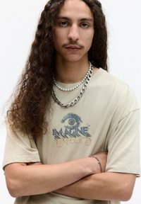 Béžové bavlnené tričko s krátkymi rukávmi, s grafickým dizajnom oka a textom "Imagine Dragons." Prekrývajúce sa strieborné a perlové náhrdelníky.