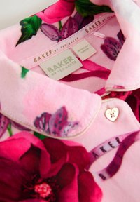 Blumenhemd mit pinkem Hintergrund, versehen mit großen roten und lila Blumen. Enthält einen goldenen herzförmigen Knopf und Etiketten mit dem Markennamen.