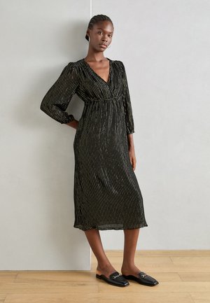 Abito nero con strisce metalliche verticali, maniche lunghe, scollatura a V, vita arricciata e lunghezza a metà polpaccio. Il modello indossa scarpe nere slip-on.