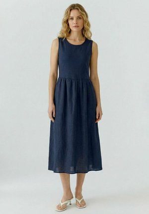 Frau mit welligem, blondem Haar, die ein ärmelloses, marineblaues Midikleid und weiße Sandalen mit Absatz trägt und vor einem schlichten hellen Hintergrund steht.