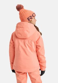 Chaqueta de nieve color melocotón con capucha y diseño texturizado, combinada con guantes negros y un gorro naranja con pompón y logo.