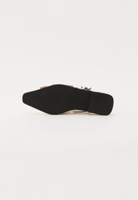 Beige høyhælte sandaler med svart såle, justerbar ankelrem og metalliske spenne detaljer. Glatt tekstur på overdelen.