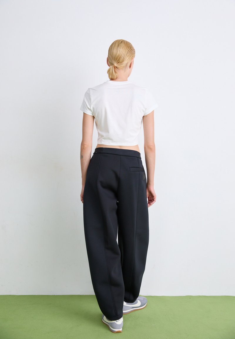 Pantaloni neri a gamba larga con una texture liscia, dotati di dettagli sulla tasca posteriore. Abbinati a una maglietta bianca aderente e corta e sneakers grigie.