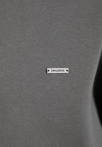 Graues Stoff-T-Shirt mit einem kleinen silbernen Metalletikett, auf dem "SMILODOX" eingraviert ist. Es zeichnet sich durch eine glatte Textur und einen figurschmeichelnden Schnitt aus.