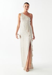 Vestido de noche de un solo hombro con lentejuelas en un tono beige claro, con detalles de volantes y una abertura en el muslo. Combinado con un bolso de mano blanco y tacones con tiras.