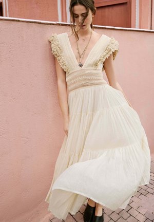 Vrouw in een vloeiende crème jurk met gestructureerde mouwen en gelaagde rok, staand tegen een roze muur, tenen zichtbaar in zwarte schoenen.