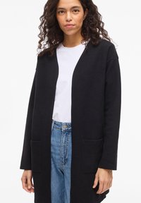 Μαύρο ζακέτα από υφασμάτινη υφή, με ανοιχτό μπροστινό μέρος, μακριά μανίκια και δύο μπροστινές τσέπες. Φορεμένο πάνω από ένα λευκό πουκάμισο και τζιν.