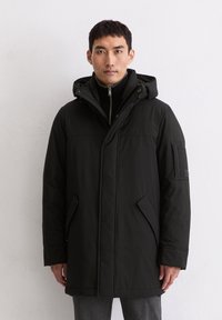 Schwarze Parkajacke mit Reißverschluss, verstellbarem Kapuzenkragen, Seitentaschen und strukturiertem Stoff. Enthält ein Abzeichen am linken Ärmel.