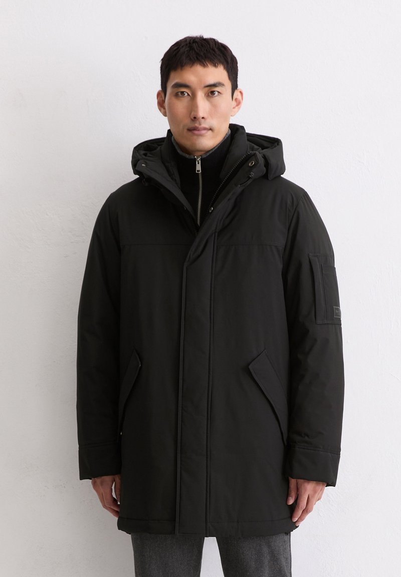 Schwarze Parkajacke mit Reißverschluss, verstellbarem Kapuzenkragen, Seitentaschen und strukturiertem Stoff. Enthält ein Abzeichen am linken Ärmel.