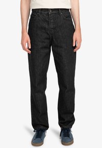 Jean en denim noir avec une coupe droite, un design à cinq poches et une fermeture à bouton. Portés avec des baskets bleues à lacets foncés et une semelle claire.