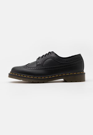 Chaussure brogue noire en cuir avec perforations en forme d'aile, lacets noirs, surpiqûres jaunes le long de la semelle et semelle en caoutchouc marron translucide.