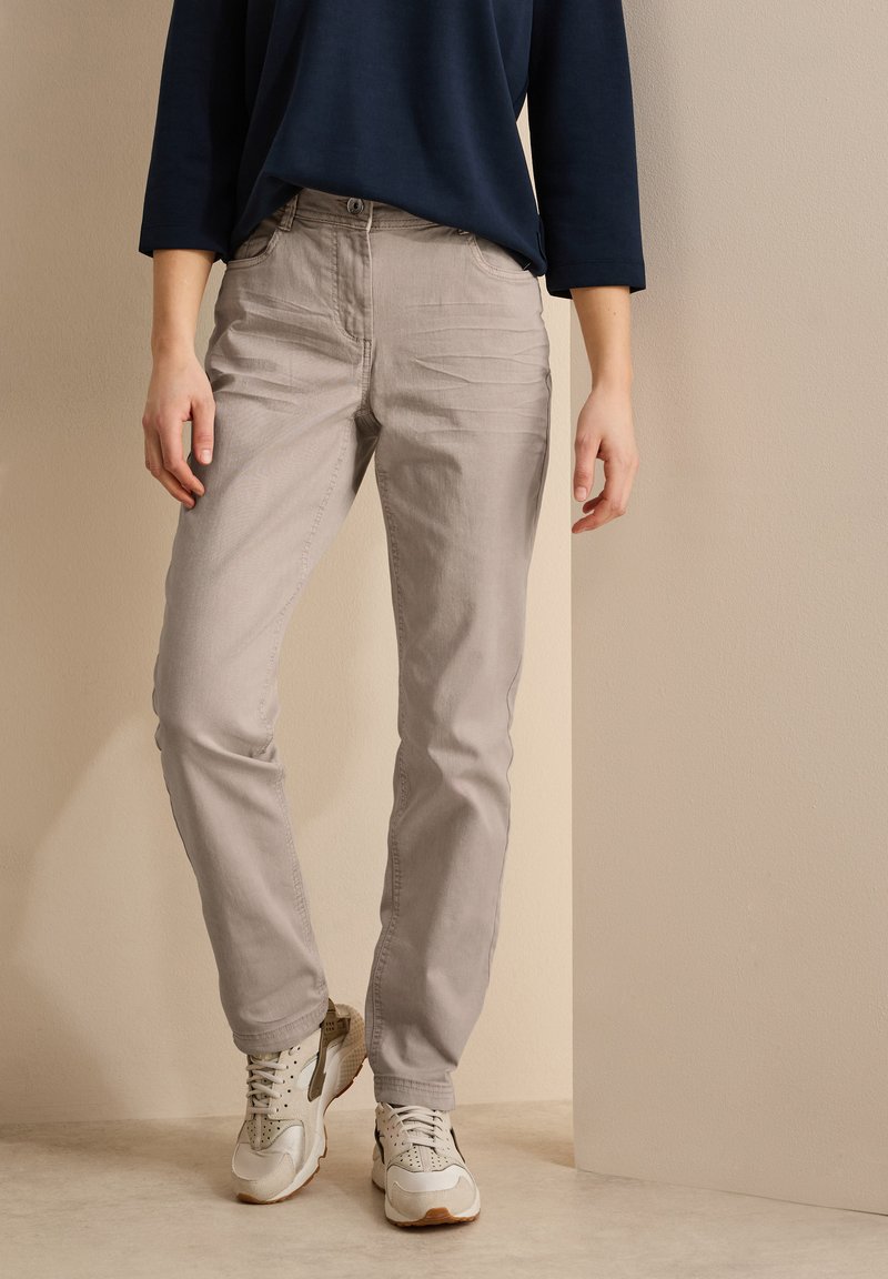 Pantalones de algodón gris claro con corte recto y bolsillos laterales, combinados con zapatillas blancas y grises, todo esto frente a un fondo neutro.