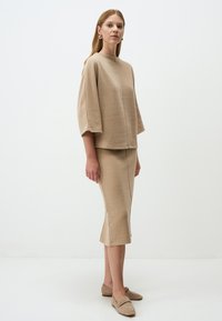 Beige Stricktop mit kurzen, breiten Ärmeln und hohem Kragen, kombiniert mit einem taillierten beige Pinselrock und passenden Slippern.