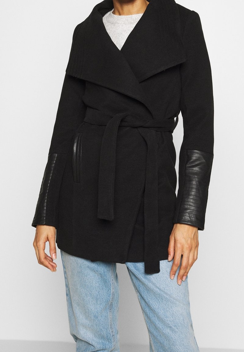 Chaqueta negra con un diseño envolvente, que presenta un cuello amplio, cinturón y mangas de cuero con un patrón texturizado. Corte casual.