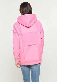 Felpa con cappuccio rosa caratterizzata da un mix di pannelli lisci e testurizzati, con un design squadrato e polsini a costine, indossata con jeans azzurri chiari.