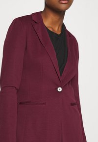 Personne portant un blazer bordeaux ajusté avec un seul bouton blanc sur un haut noir, montré de la poitrine au menton.