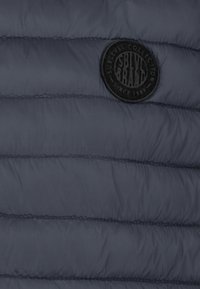 Dunkelblauer gestepptes Gewebe mit horizontalen Nähten und einem runden schwarzen Aufnäher mit der Aufschrift "SBLVYS BRAND" in der oberen rechten Ecke.