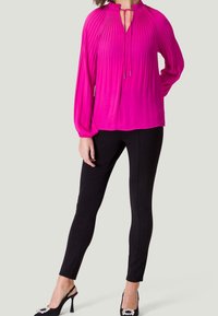 Blouse fuchsia à manches longues avec des plis verticaux, col mandarin et détail à nouer. Associée à des leggings noirs ajustés et des sandales à talons noires.