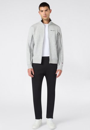 Junger Mann trägt eine hellgraue Zip-up-Jacke, ein weißes T-Shirt, schwarze Hose und weiße Turnschuhe und steht vor einem schlichten hellen Hintergrund.