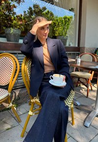 Marinblazer över en beige tröja, vida mörka jeans, håller i en kaffekopp på en caféterass med flätade stolar och soligt väder.