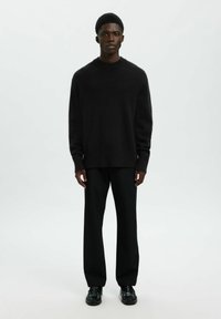 Joven de pie, vistiendo un suéter negro de manga larga, pantalones rectos negros y zapatos de vestir negros sobre un fondo claro y liso.