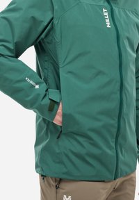Veste imperméable verte avec une fermeture éclair intégrale, en tissu texturé, avec des poignets réglables et un détail de logo. Elle possède une poche latérale et un cordon de serrage à l'ourlet.