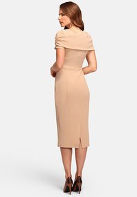 Woman with long brown hair wearing a fitted beige midi dress with ruched sleeves and a back slit, paired with dark brown high heels.