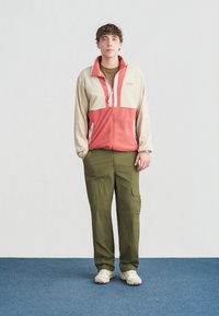 Άνδρας που στέκεται σε μπλε χαλί, φοράει μπεζ και κοραλλί φλις με φερμουάρ, χακί παντελόνι καμπαρντίνα και λευκά αθλητικά παπούτσια, με τα χέρια στις τσέπες του, ουδέτερη έκφραση.