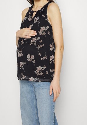 Kobieta w ciąży w czarnej, kwiecistej bluzce bez rękawów i jasnoniebieskich dżinsach, delikatnie trzymająca brzuch jedną ręką.