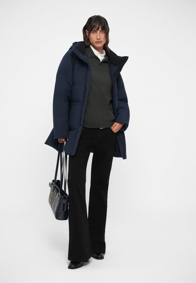 Doudoune marine avec un col haut, portée sur un pull foncé et un pantalon noir évasé. Un sac à main noir complète la tenue.