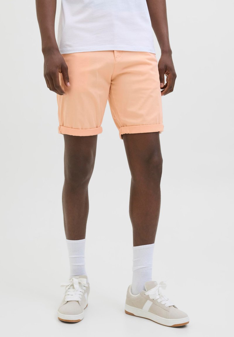 Uomo che indossa pantaloncini color pesca con orlo arrotolato, calzini bianchi, scarpe da ginnastica beige e camicia bianca, in piedi su uno sfondo chiaro e uniforme.