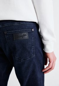 Jeans de mezclilla azul oscuro con un parche de cuero Wrangler en el bolsillo trasero, con remaches plateados y un diseño clásico de cinco bolsillos.