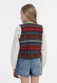 Bunt gestrickte Weste mit rotem Grund, die mehrfarbige horizontale Streifen und dekorative Muster aufweist. Kombiniert mit einem weißen strukturierten Oberteil und Jeansshorts.