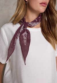Mørkerød bandana-halstørklæde med hvidt paisley-mønster, bundet om halsen, båret over en simpel hvid t-shirt. Stoffet ser let og blødt ud.