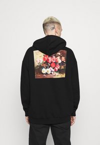 Sudadera negra con una ilustración impresa de flores en la parte posterior, con vibrantes flores rojas, rosas y blancas sobre un fondo atenuado.