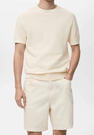 Mężczyzna w kremowym, teksturowanym swetrze z krótkim rękawem i do kompletu kremowych, sięgających kolan szortach, stojący z jedną ręką w kieszeni.