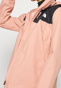 Veste imperméable rose clair avec une section supérieure noire, dotée d'une fermeture éclair, d'une capuche ajustable et d'un logo sur la poitrine.